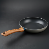 A minimalist aluminum frying pan with a curved wooden handle, photographed on a dark gray studio background.