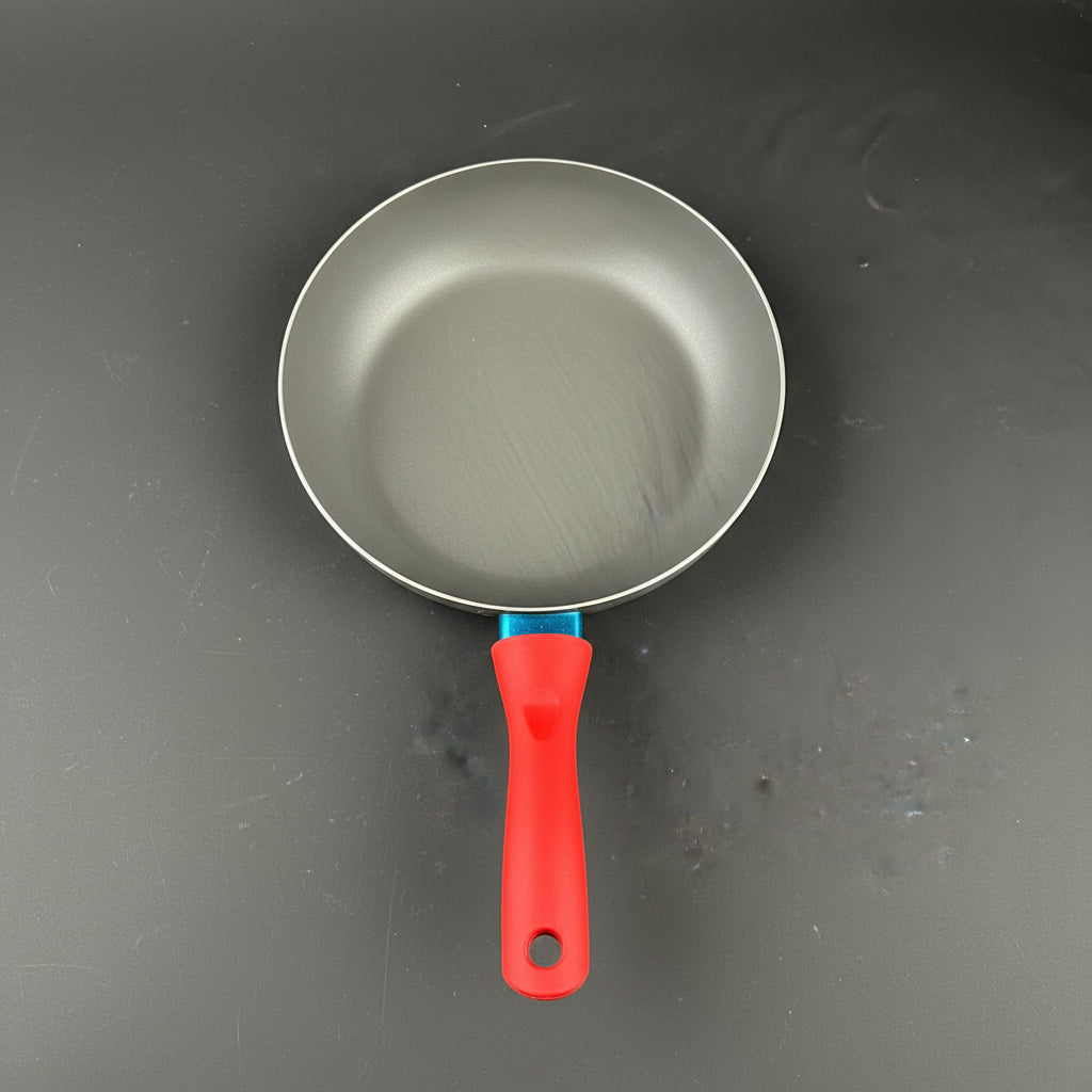 A close-up of a frying pan handle with a bright red ergonomic grip and a metallic silver pan body, resting on a dark gray surface.