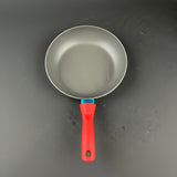 A close-up of a frying pan handle with a bright red ergonomic grip and a metallic silver pan body, resting on a dark gray surface.