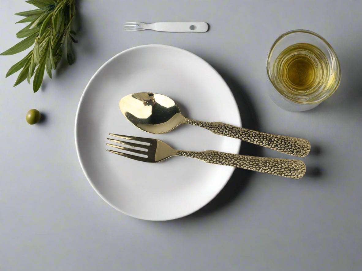 Gold cutlery set on a white plate with a glass of white wine and greenery on a light gray background